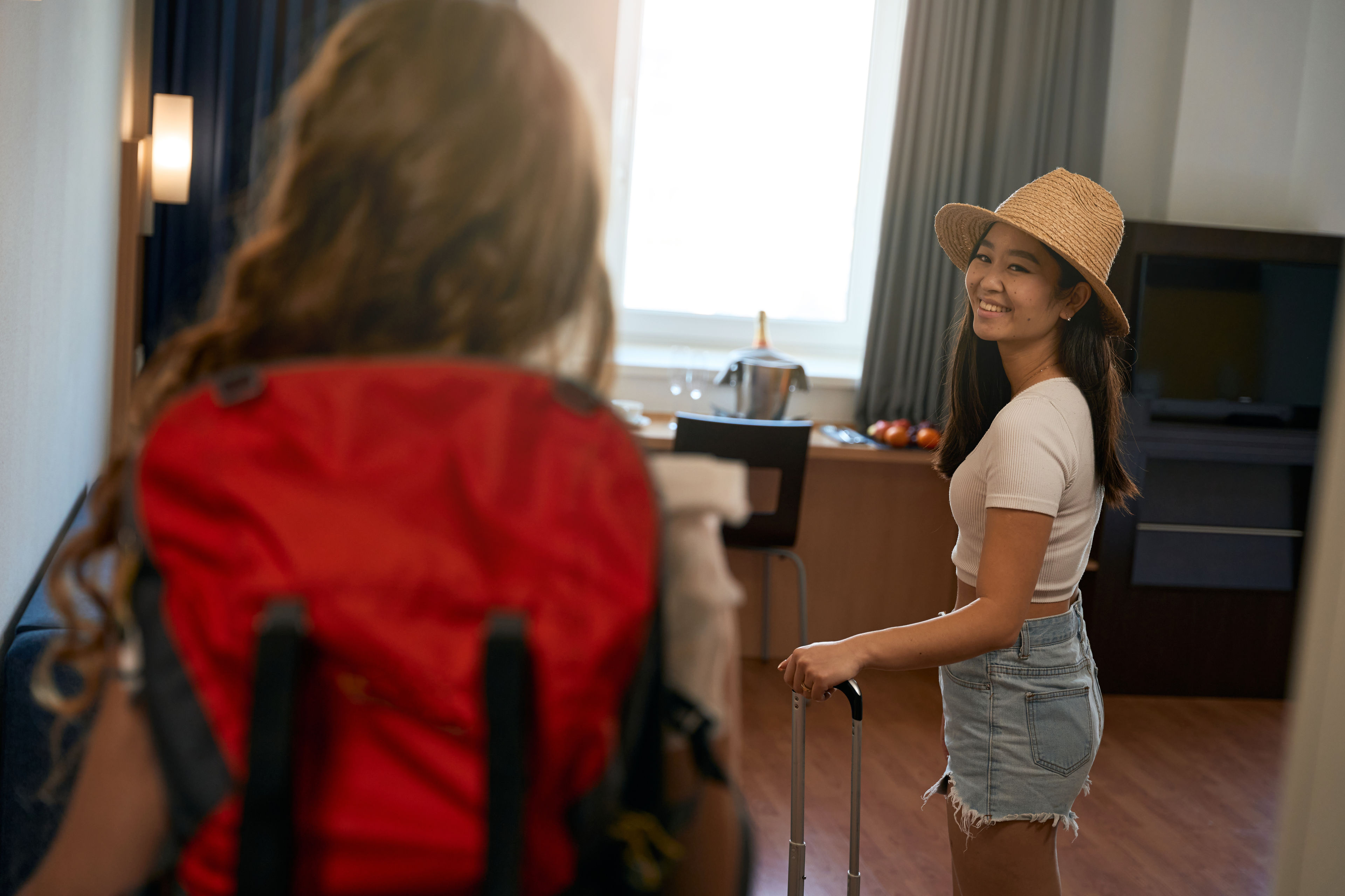A smiling woman wearing a hat stands inside a hotel room 