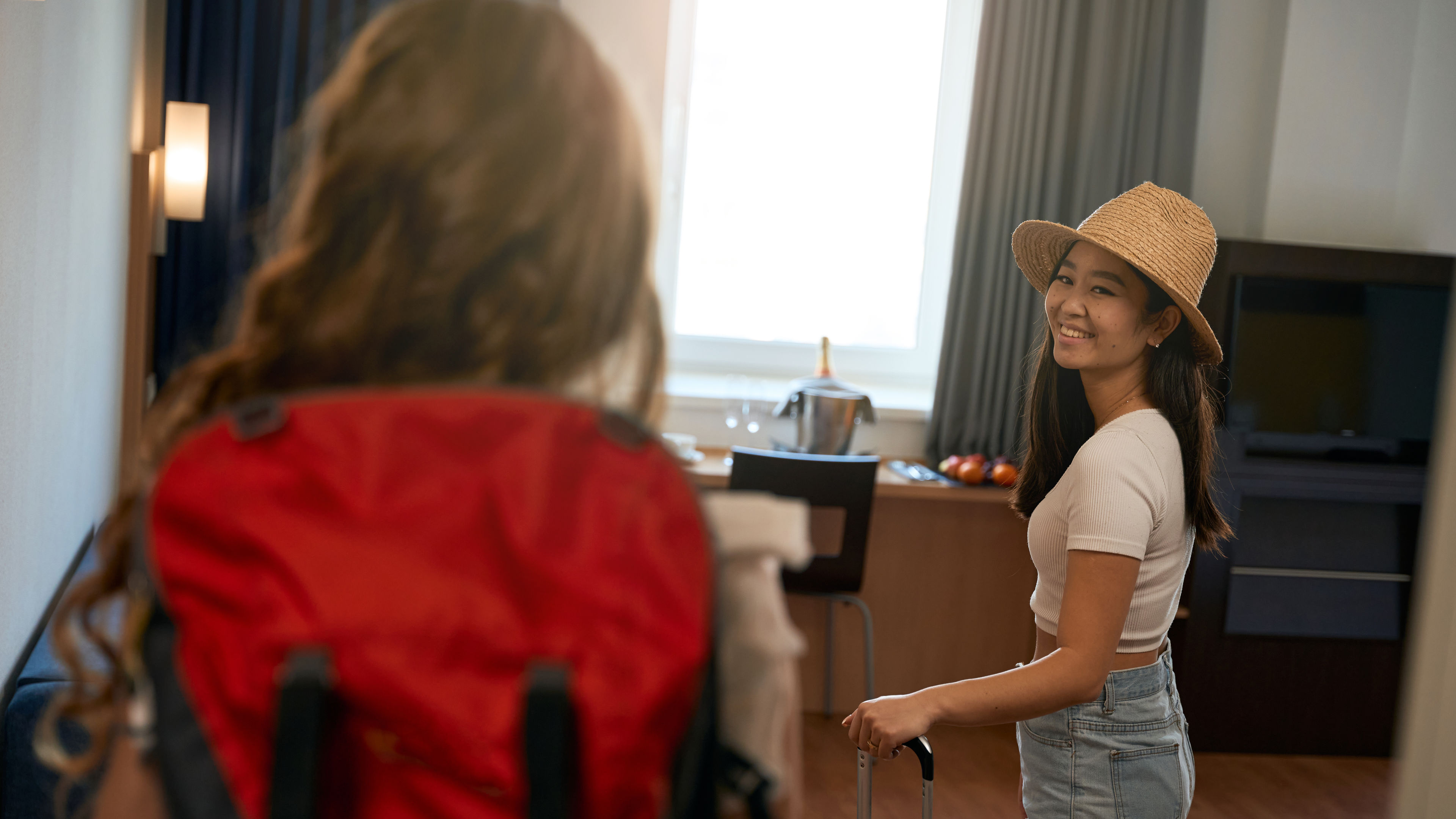 A smiling woman wearing a hat stands inside a hotel room 